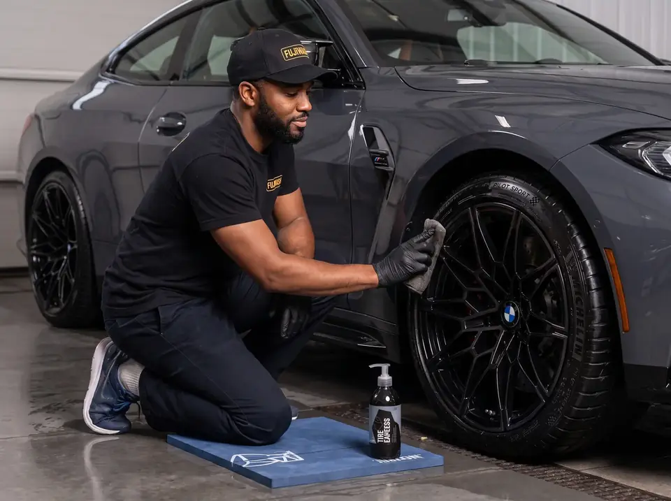 FujiWash™ detailer in a branded black cap and shirt kneeling beside a grey BMW coupe, dressing the black alloy wheel with a microfiber towel during an interior and exterior detail.